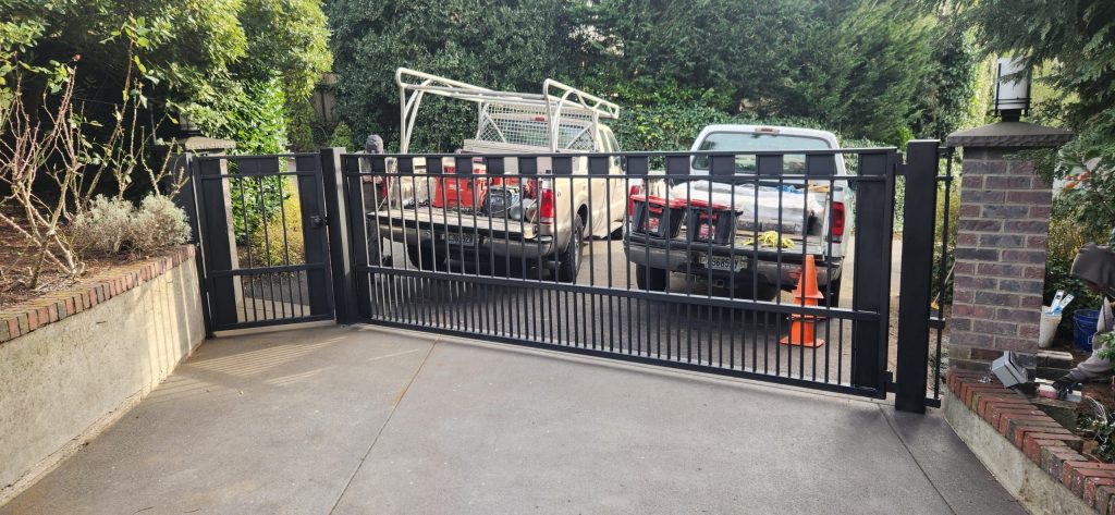 A black metal sliding gate partially open in front of a driveway with two parked pickup trucks and orange cones.