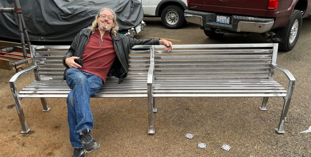 A man sits and smiles on a large metal bench in a parking lot, holding a phone with cars in the background.