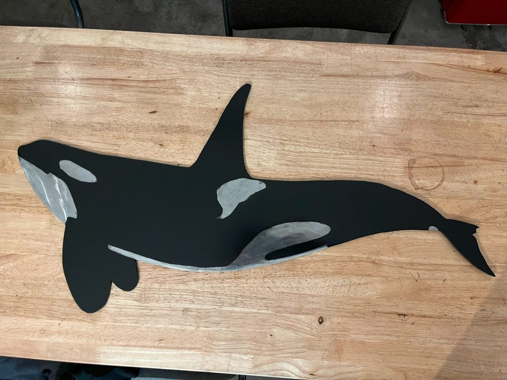 A flat, black and white painted cutout of an orca whale lies on a wooden table.