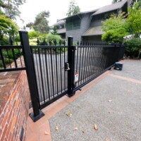 Black metal security gate at the entrance of a driveway leading to a house with trees and shrubs in the background.