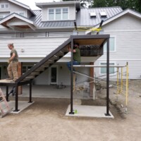 Three people work on installing a metal and wood staircase structure outside a two-story house under construction.