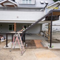 Two workers build a metal staircase frame outside a modern house, with tools and scaffolding nearby.