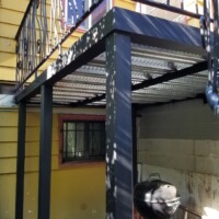 A metal porch with decorative railing is supported by four steel posts above a small outdoor area with a grill.