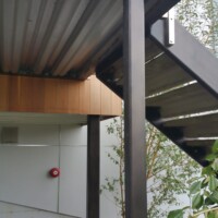 A metal staircase with black supports and a corrugated underside is shown next to a modern building exterior and some plants.