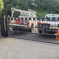 A black metal sliding gate partially open in front of a driveway with two parked pickup trucks and orange cones.